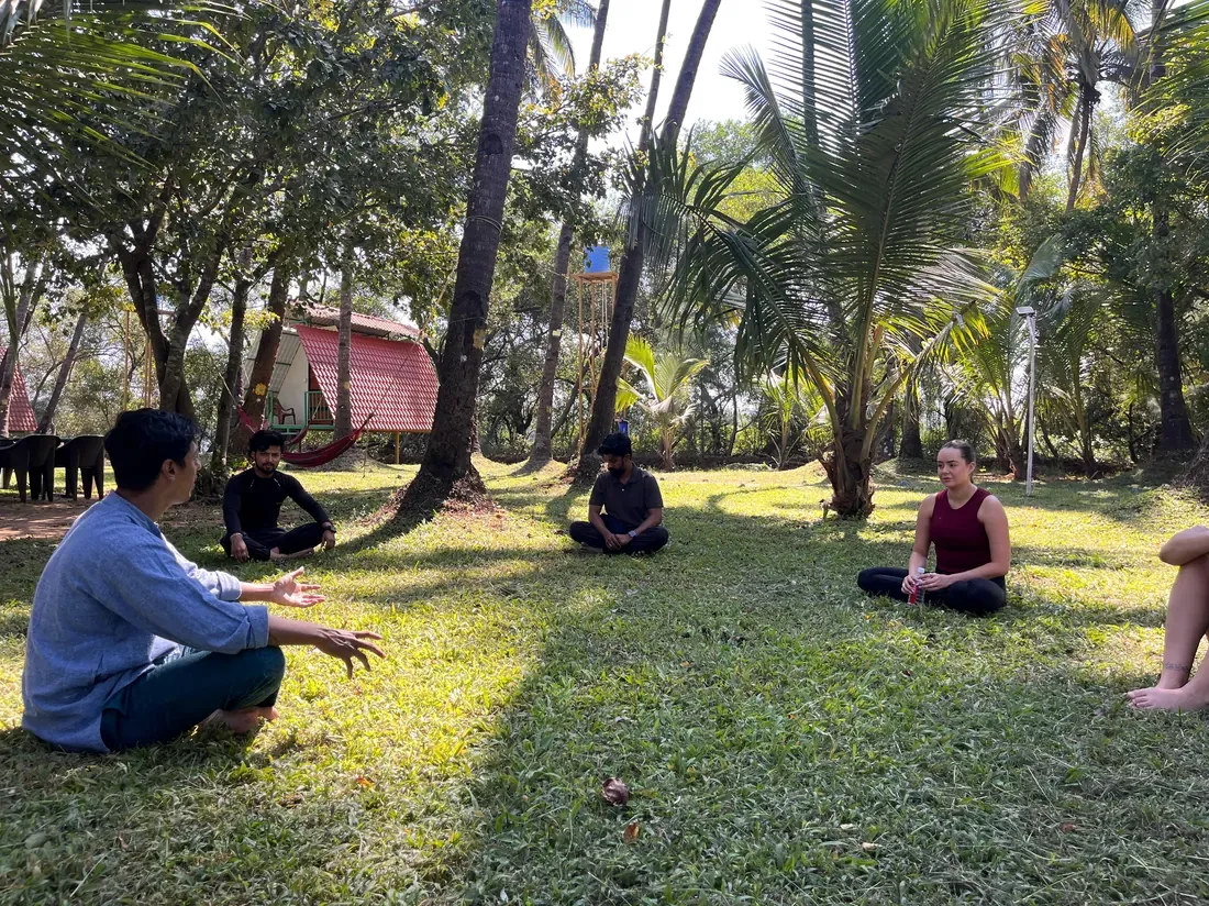 A facilitator speaking with a small group seated on the grass among the palms