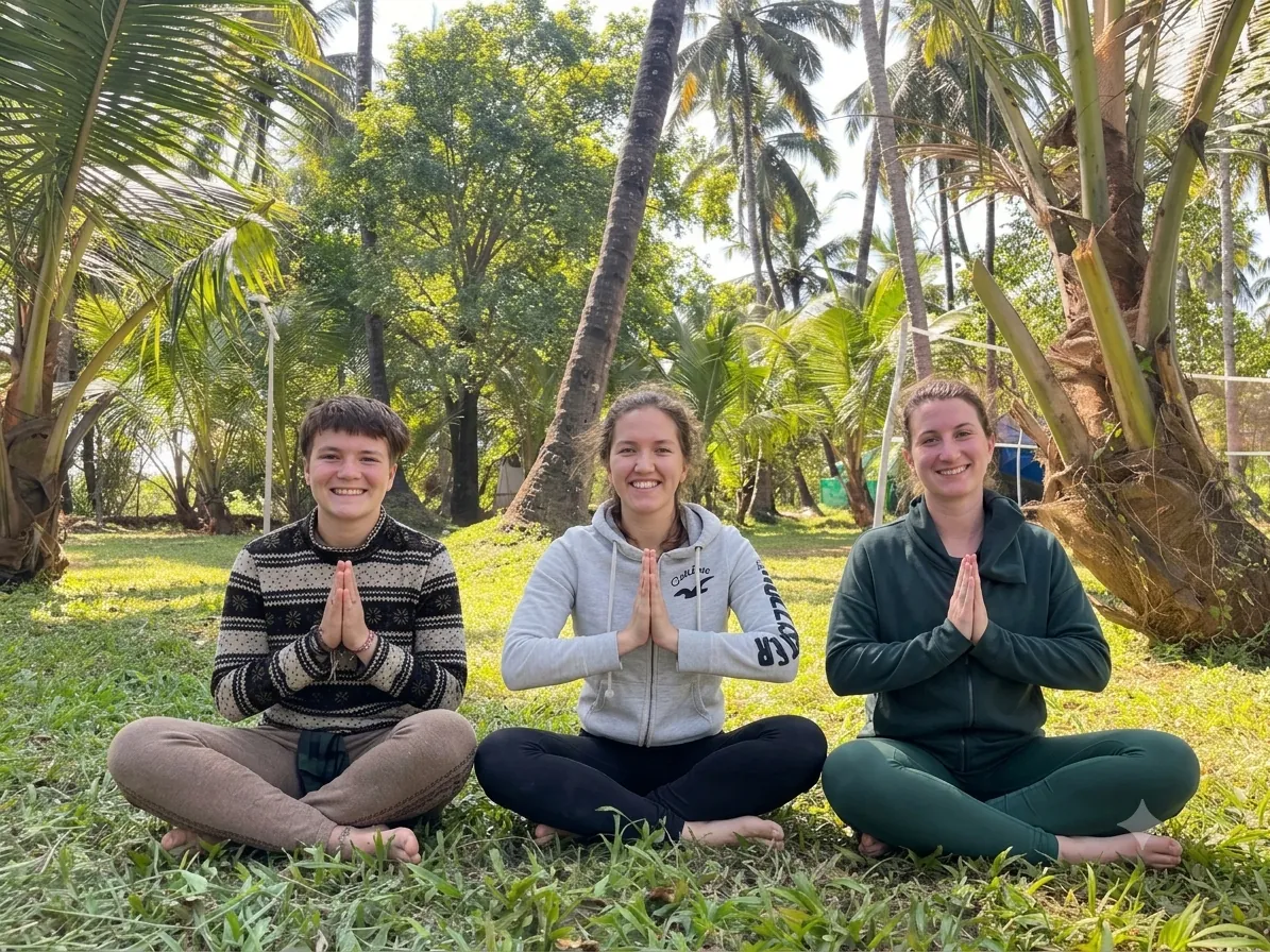 Three smiling guests seated on the grass with hands in namaste